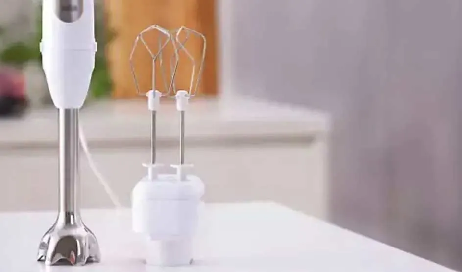 Een witte staafmixer en kloppers staan op een wit aanrechtblad. Achtergrond is een keuken.