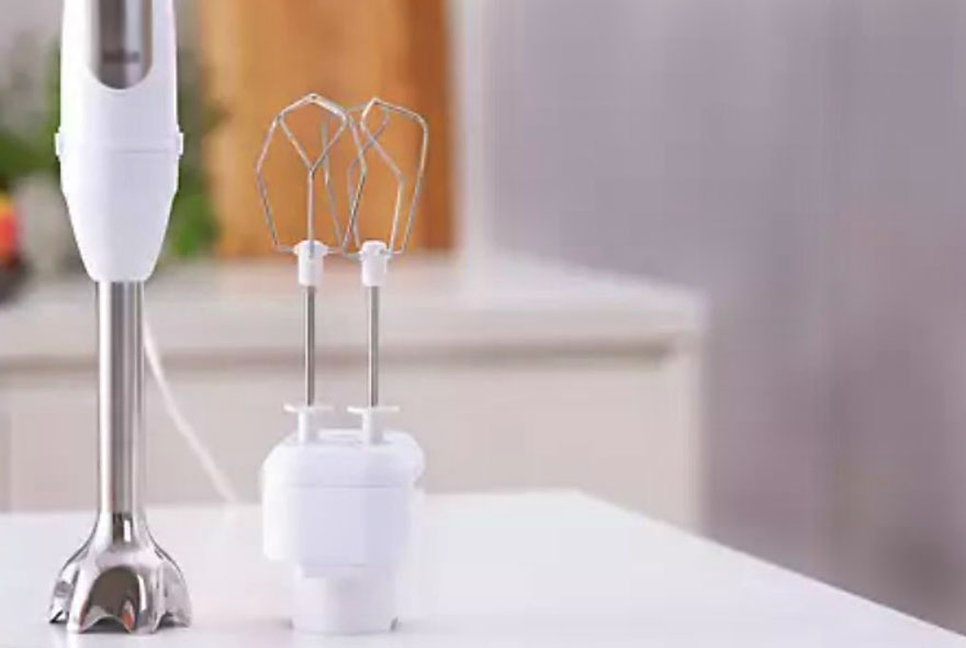 Een witte staafmixer en kloppers staan op een wit aanrechtblad. Achtergrond is een keuken.