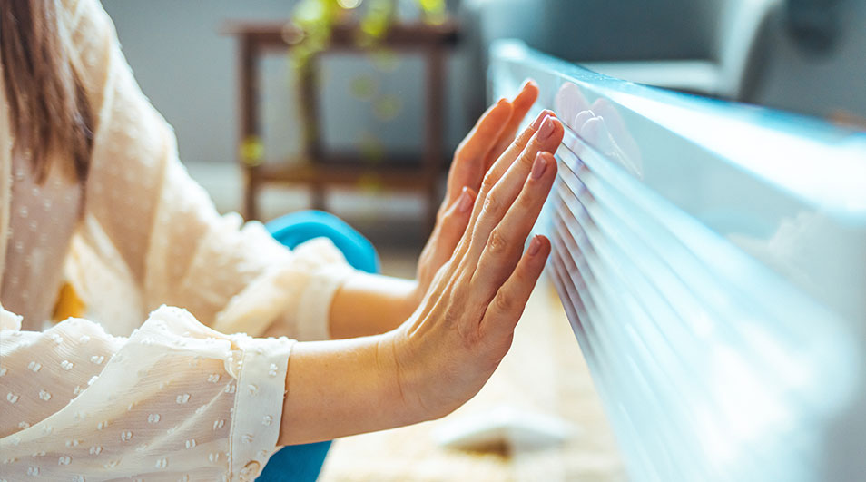 Twee handen duwen tegen een witte, geplooide jaloezie. Achtergrond: wazige kamer met een houten tafel en een blauwe stoel.