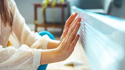 Une personne en chemisier blanc appuie les mains contre un store blanc. Une chaise bleue et une table en bois sont en arrière-plan.