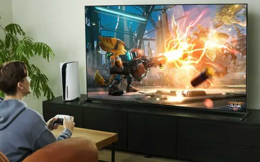 Un joueur joue à Ratchet & Clank sur un grand écran assis sur un canapé tenant une manette blanche, avec une PlayStation 5 et des plantes vertes visibles dans la pièce.