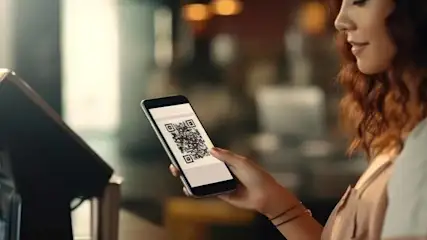 Une personne tient un smartphone affichant un code QR près d'un terminal de paiement noir. L'arrière-plan est flou.