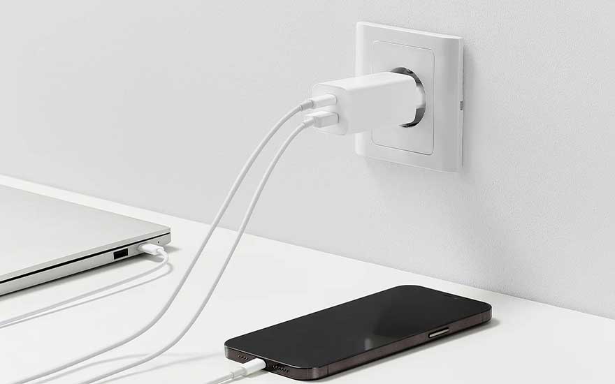 Un chargeur blanc est branché sur une prise murale blanche. Un ordinateur portable et un téléphone noir sont en charge via des câbles blancs.