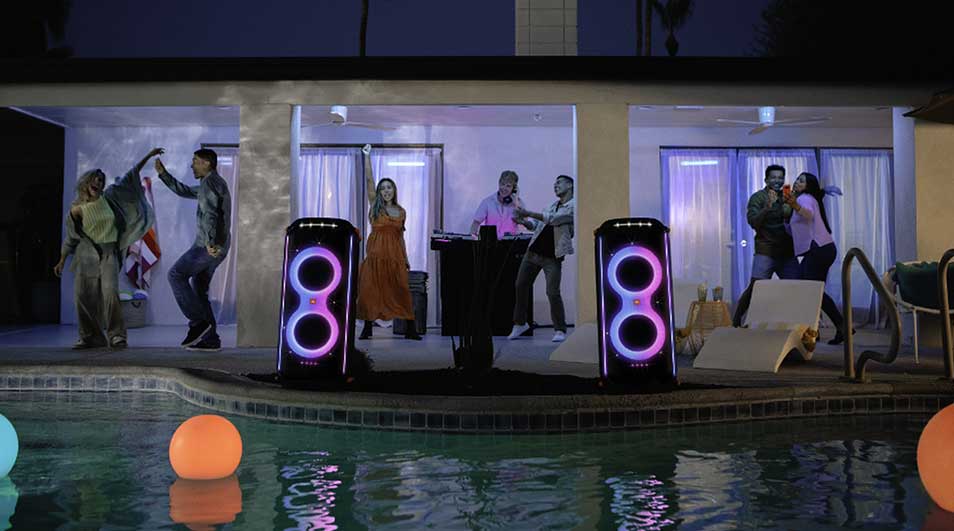 Two speakers, lit with purple circles, flank a DJ setup near a pool with floating orange and blue balls. The background shows a house.
