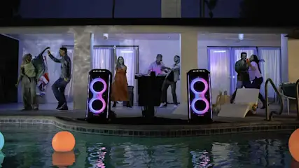 Fête nocturne au bord de la piscine avec lumières néon, haut-parleurs éclairés, eau reflétant la lumière et boules flottantes colorées.