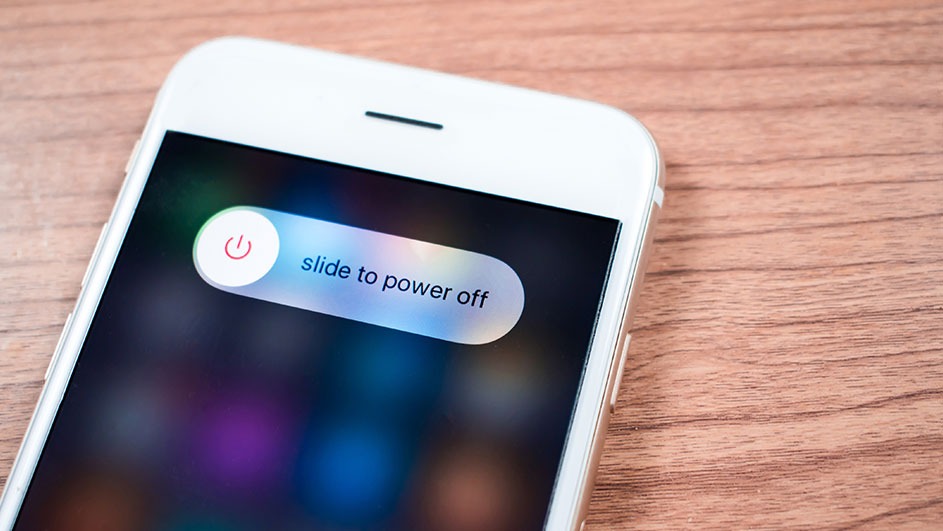 Un iPhone blanc affiche un curseur "slide to power off" sur un fond d'écran sombre et coloré, sur une table en bois.