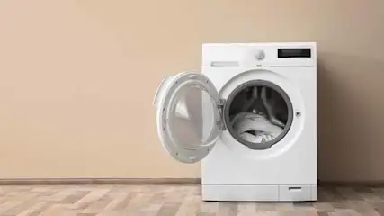 Une machine à laver blanche avec la porte ouverte dans une pièce aux murs beiges et au parquet.