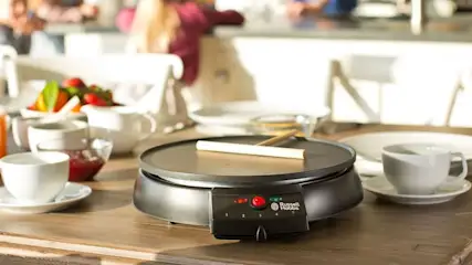 Table en bois avec crêpière noire Russell Hobbs, spatule blanche, vaisselle blanche et fruits.