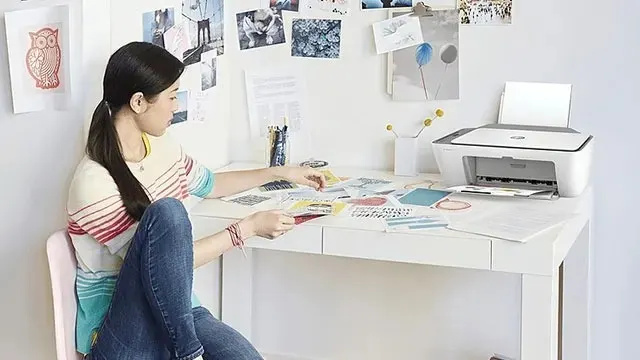 Een witte printer staat op een bureau vol papieren en kantoorbenodigdheden, tegen een witte muur met foto's.