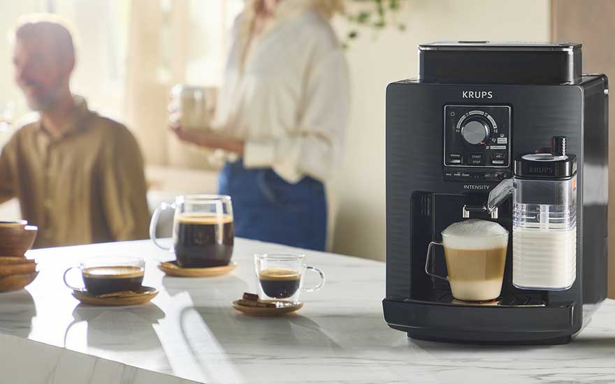Krups-koffiemachine bereidt een romige latte op een marmeren tafel, met pratende mensen sfeervol onscherp op de achtergrond.