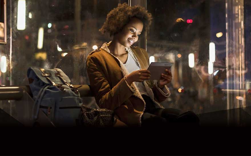 Een blauwe rugzak staat naast iemand die een tablet gebruikt in een bushokje. Buiten zijn stadslichten zichtbaar.