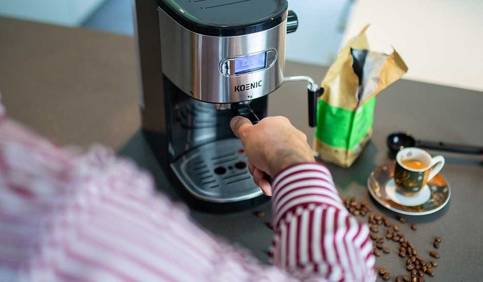 A person in a striped shirt operates a KOENIC coffee machine on a counter scattered with beans, beside an open bag and a tiny espresso cup.