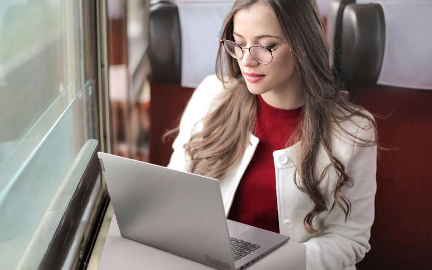 Une femme portant des lunettes utilise un ordinateur portable argenté dans un train, tandis que le paysage défile derrière la grande fenêtre.