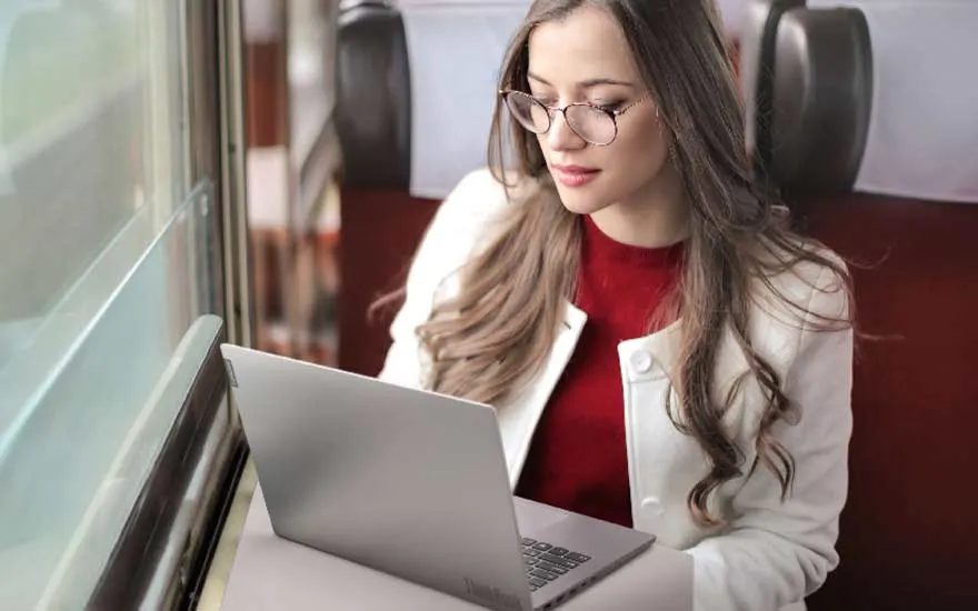 Une femme portant des lunettes utilise un ordinateur portable argenté dans un train, tandis que le paysage défile derrière la grande fenêtre.