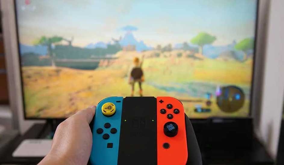 Une main tient une Nintendo Switch avec des Joy-Con bleu et rouge, affichant sur la télé le jeu The Legend of Zelda: Breath of the Wild.