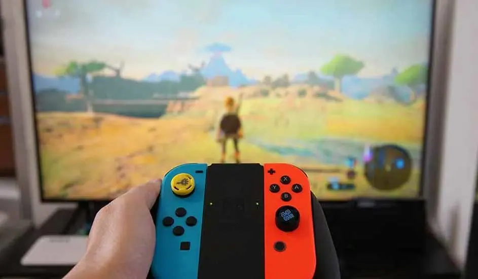 Een hand houdt een Nintendo Switch met blauwe en rode Joy-Cons, terwijl op de tv het spel The Legend of Zelda: Breath of the Wild speelt.