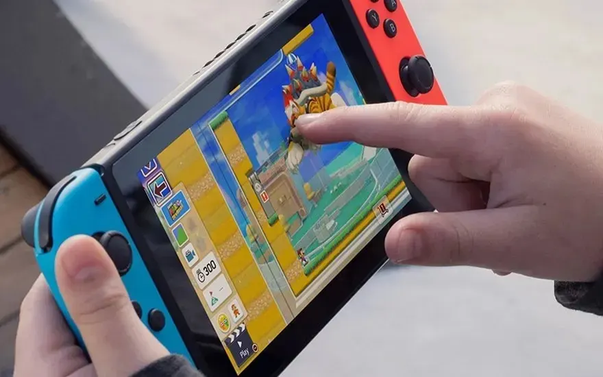 Hands hold a Nintendo Switch while a finger drags a Mario enemy across the colourful level editor on the bright touchscreen