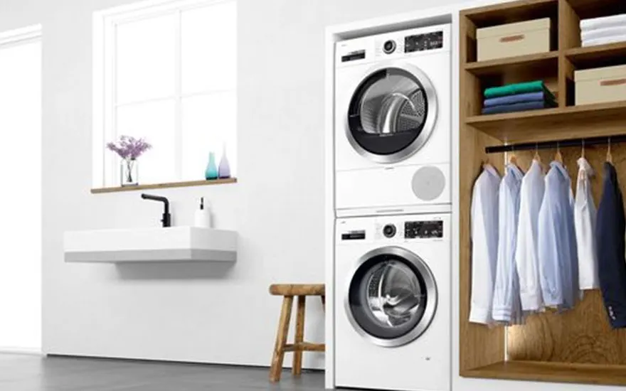 A laundry room features a stacked white washer and dryer, a sink with a black faucet, and a wooden shelf unit with clothes.