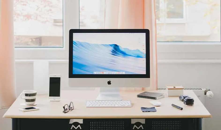 Apple iMac with keyboard on a desk, iPhone and accessories around it; clear Apple logo visible on iMac and iPhone.