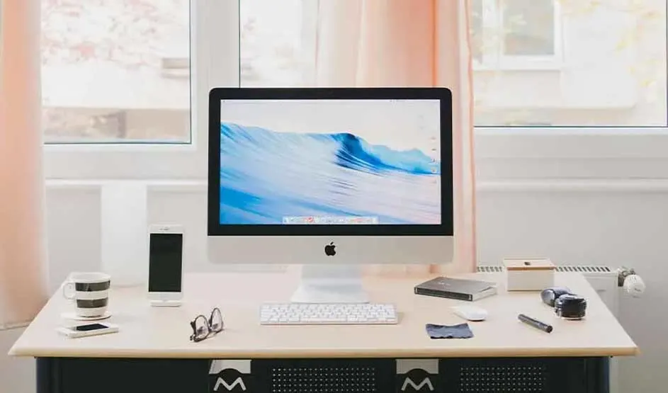 Apple iMac with keyboard on a desk, iPhone and accessories around it; clear Apple logo visible on iMac and iPhone.