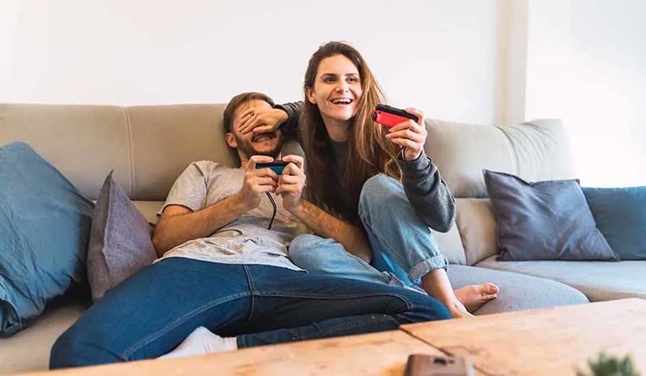 Twee mensen zitten ontspannen op een bank en spelen samen een videogame met Nintendo Switch-controllers in een gezellige woonkamer.