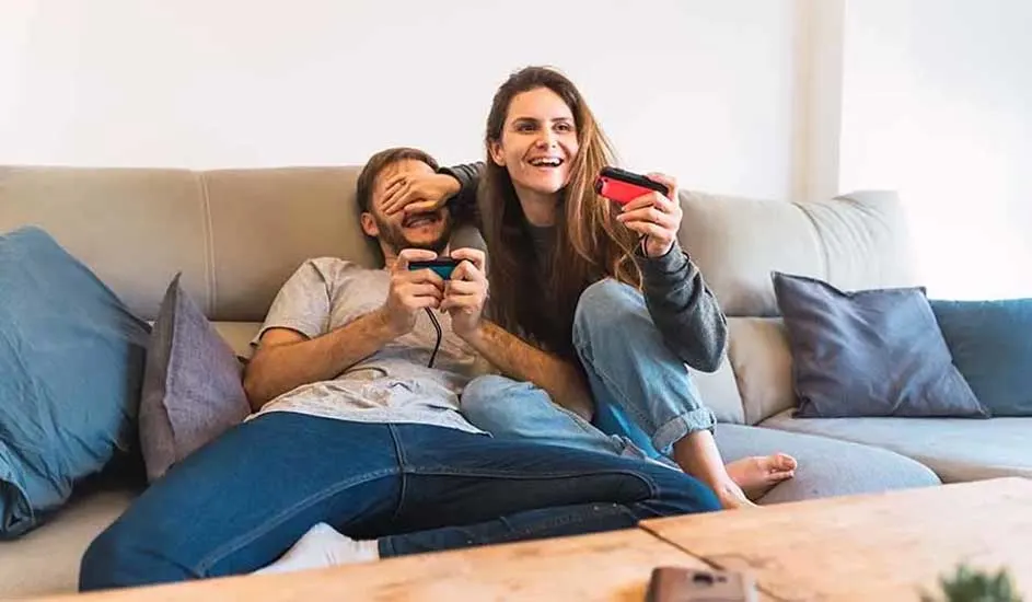 Deux personnes sont assises sur un canapé, jouant ensemble à un jeu vidéo avec des manettes Nintendo Switch, dans un salon accueillant.