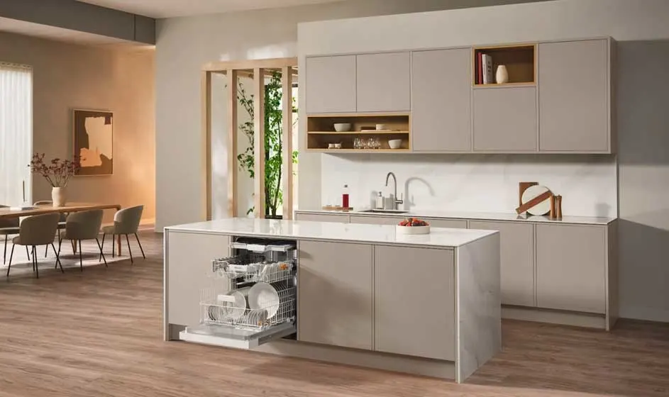 Une cuisine moderne avec armoires blanches, un îlot avec lave-vaisselle intégré et accents en bois. L'espace s'ouvre sur une salle à manger et un salon avec lumière naturelle. Design minimaliste avec sol en bois et solutions de rangement intégrées.