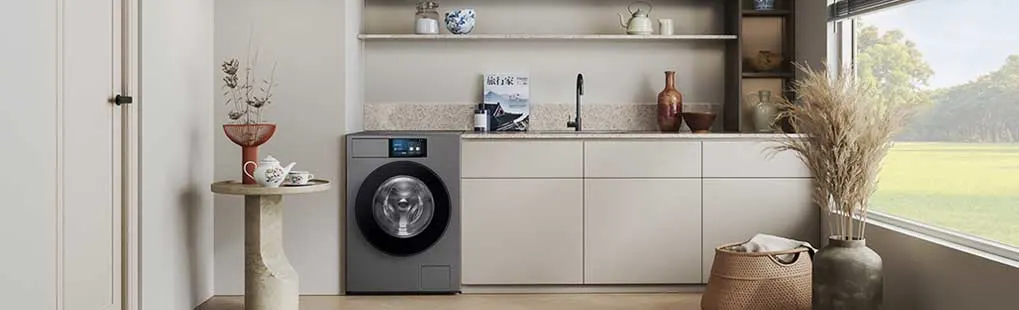 Strakke wasruimte met grijze kasten, open nis met decor, wastafel en zwarte kraan, wasmachine, hanglamp en groot raam met groen uitzicht.