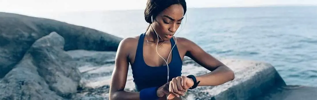 A person in sportswear checks a smartwatch on a rocky coast near the sea, with headphones in.