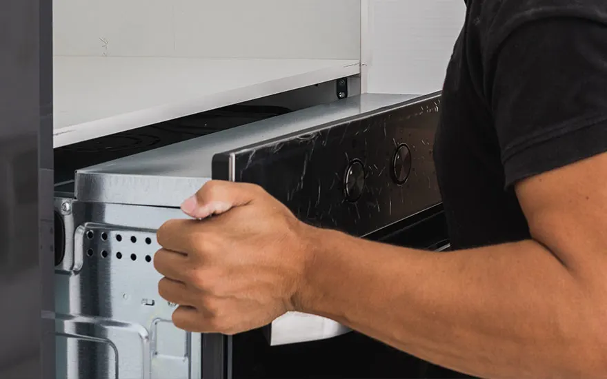 Een hand trekt een zilverkleurige oven uit een kast. De oven heeft een zwart bedieningspaneel met twee ronde knoppen.