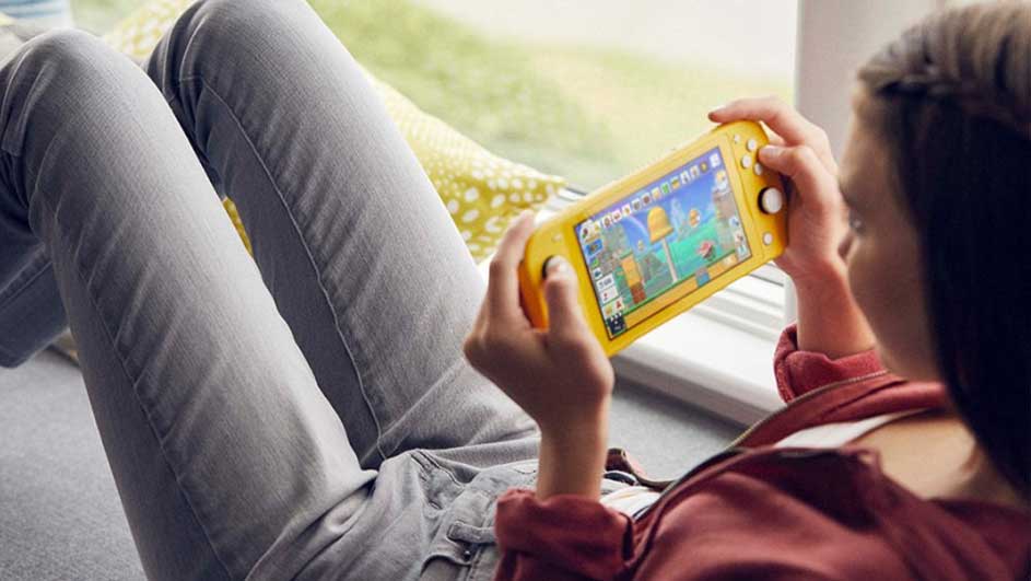 Une console jaune affiche un jeu vidéo coloré avec des arbres. Une personne assise porte un pantalon gris et une veste bordeaux.