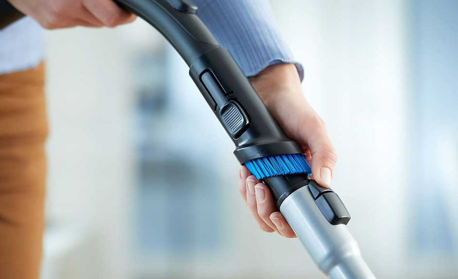 Une main ajuste une brosse bleue sur un tube d'aspirateur noir et gris. Arrière-plan flou en blanc et marron.
