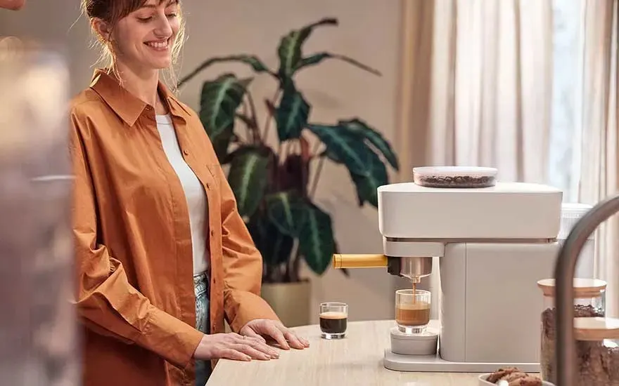 Vrouw in oranjebruin hemd glimlacht aan aanrecht terwijl witte koffiemachine vers laagjeskoffie in glas schenkt.