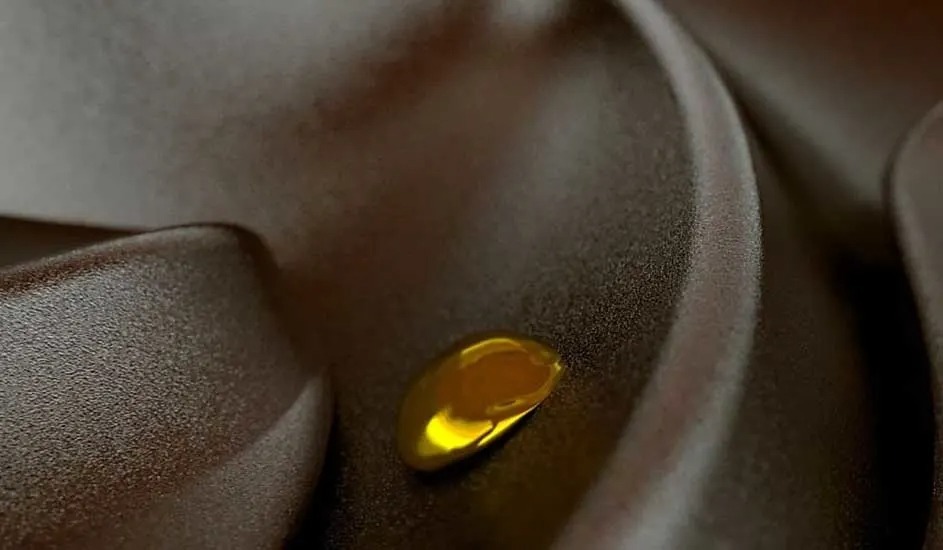 A glossy golden drop of oil rests on soft beige fabric with flowing folds in warm, muted light.