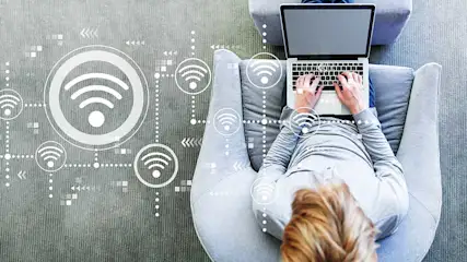 Une personne assise dans un fauteuil gris utilise un ordinateur portable, avec des symboles WiFi autour.