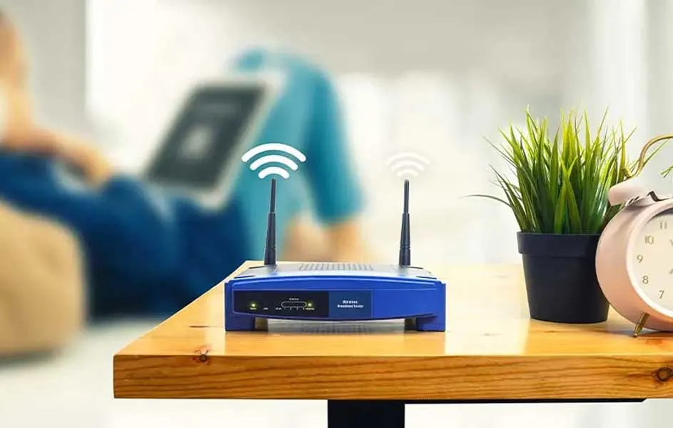 Un routeur bleu avec deux antennes sur une table en bois, à côté d'une plante et d'un réveil rose. Arrière-plan flou.