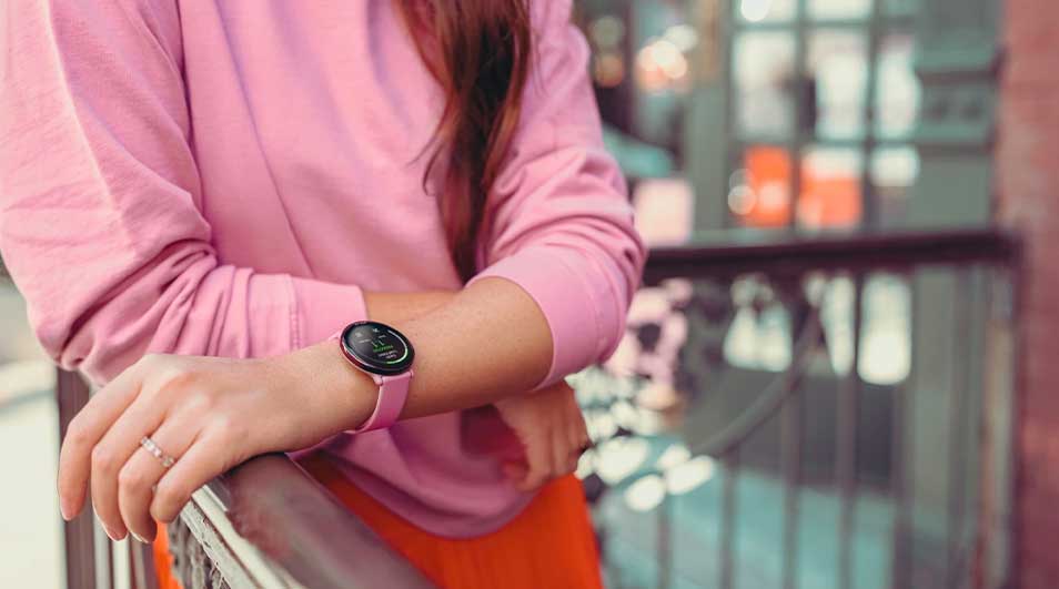 A woman wears a pink smartwatch while standing outdoors and checking her wrist, with a modern urban setting visible behind her.