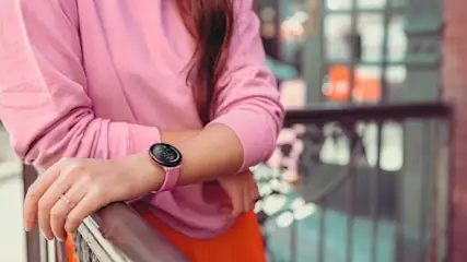 Une femme porte une montre connectée rose et consulte son poignet, tandis qu’un environnement urbain moderne apparaît derrière elle.