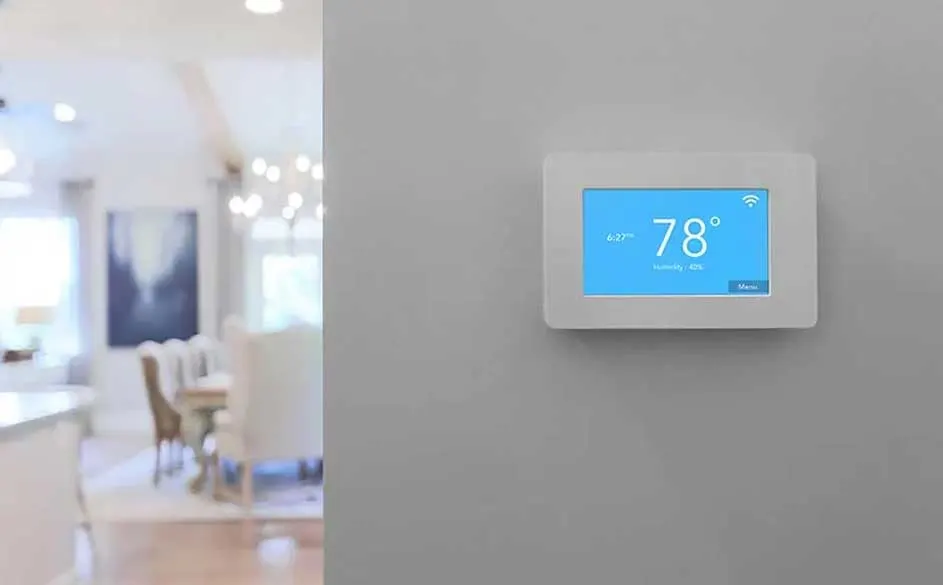 Witte thermostaat aan grijze muur toont 78 graden op een blauw scherm. Op de achtergrond is een onscherpe eetkamer te zien.