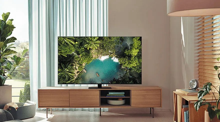 A Samsung TV displays a vivid image of a tropical lake surrounded by green trees in a stylish, bright living room with wooden furniture.