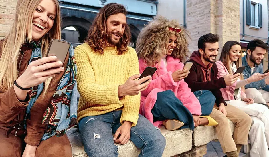 Des personnes sont assises sur un mur de pierre et regardent des smartphones. Les vêtements sont de couleurs variées.