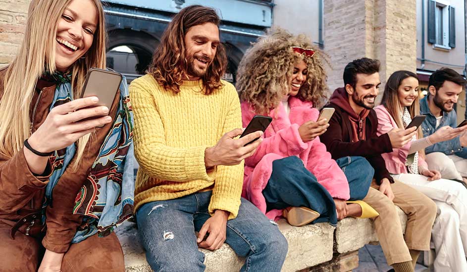 Een groep mensen zit op een stenen muur en kijkt naar hun telefoons. Ze dragen kleurrijke kleding.