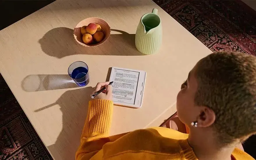 Een tafel met een e-reader, pen, waterkan, glas water en een kom met fruit. Iemand schrijft op de e-reader.