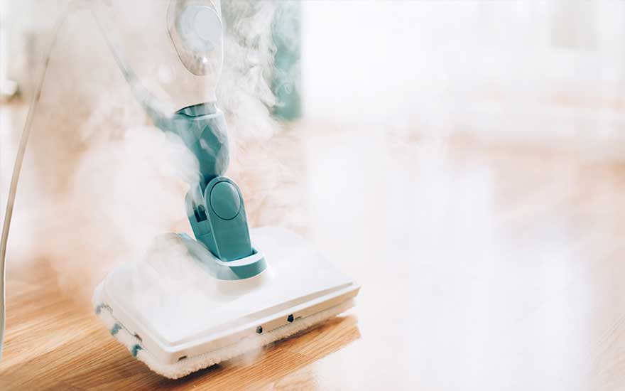 Un nettoyeur vapeur blanc et turquoise est utilisé sur un plancher en bois clair, avec de la vapeur visible.