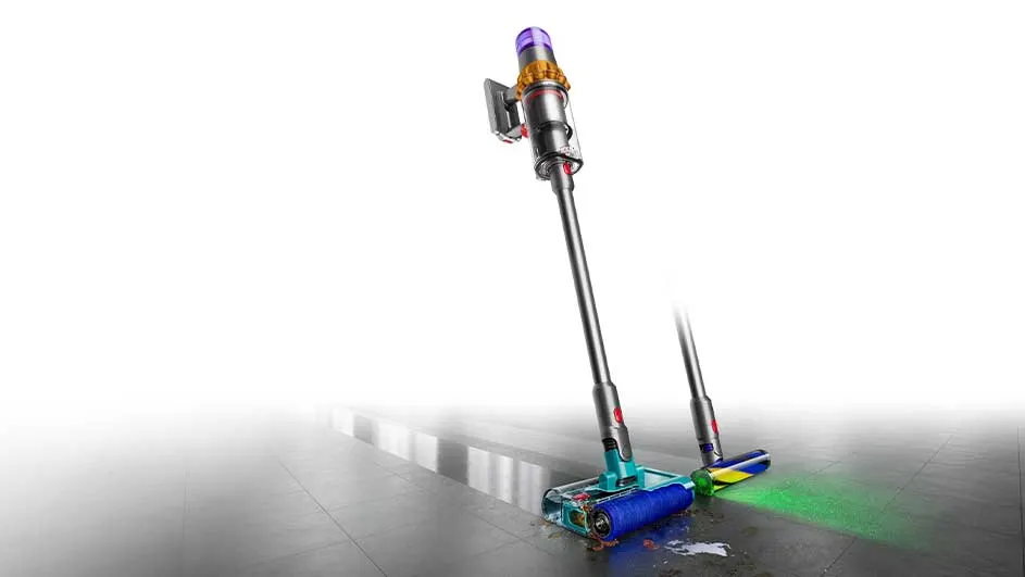 Un aspirateur-balai gris avec un collecteur violet nettoie un sol carrelé gris. La brosse est bleue et verte.