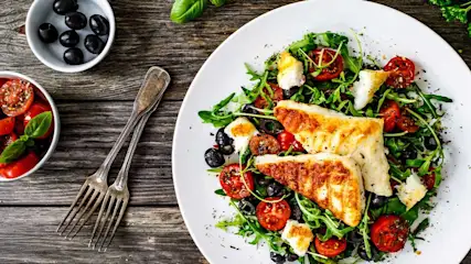 Salade avec poisson grillé, tomates, olives noires, roquette et fromage sur assiette blanche. Fourchettes à côté, sur table en bois.