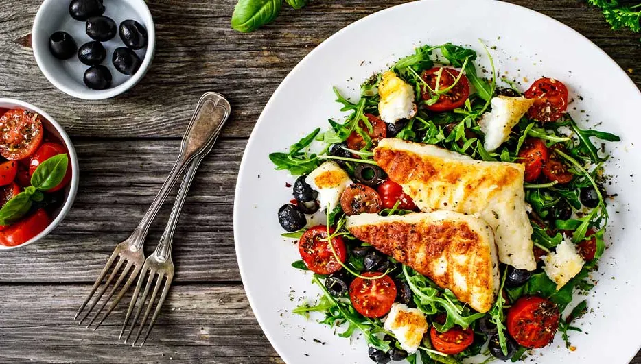 Salade avec poisson grillé, tomates, olives noires, roquette et fromage sur assiette blanche. Fourchettes à côté, sur table en bois.