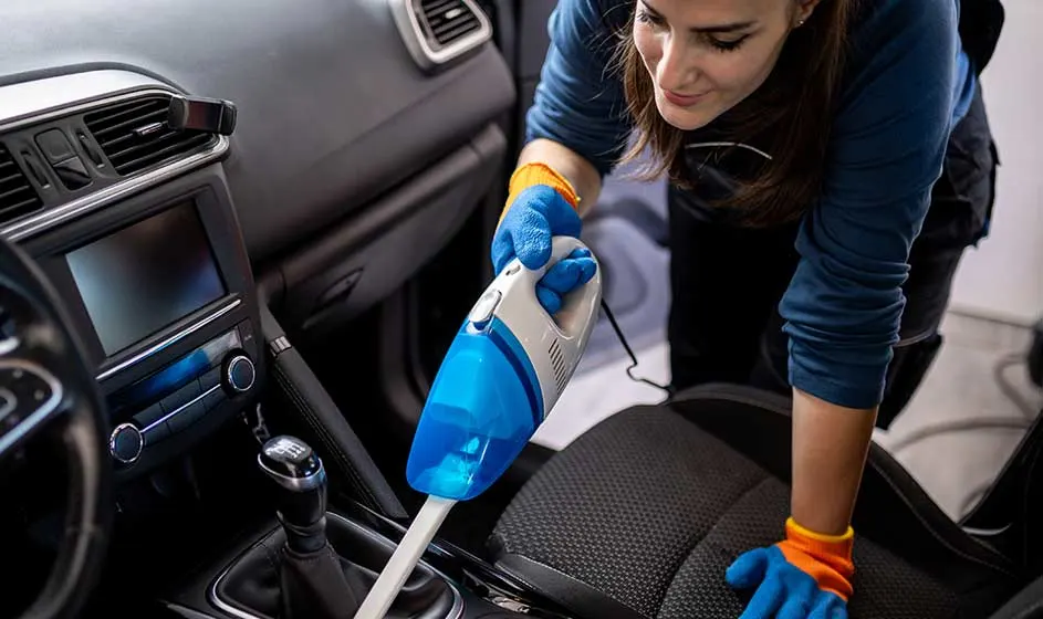 Une femme aspire soigneusement l’intérieur d’une voiture ; le tableau de bord, le siège et la console centrale sont bien visibles.