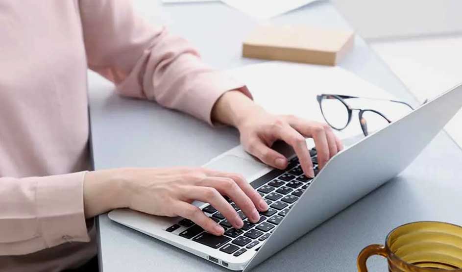 Person in pink blouse types on an Apple MacBook; glasses lie nearby on table, Apple logo visible, yellow mug in foreground
