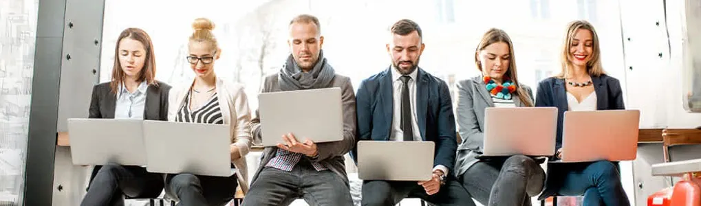 Five people are sitting side by side, each holding laptops. The laptops are silver. The background is a bright room with a large window.